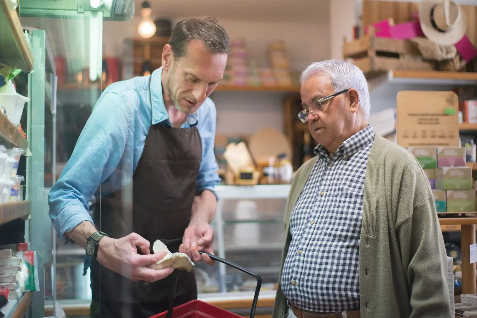 A shop worker in an apron helps an older man with an item at a grocery counter