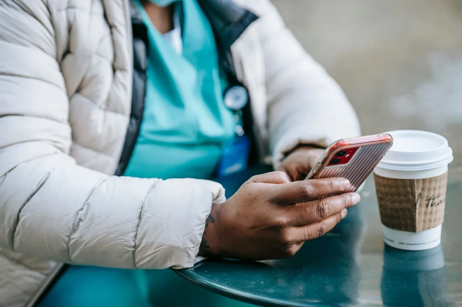 A nurse in scrubs and a puffer jacket reads her phone at a table beside a takeaway coffee