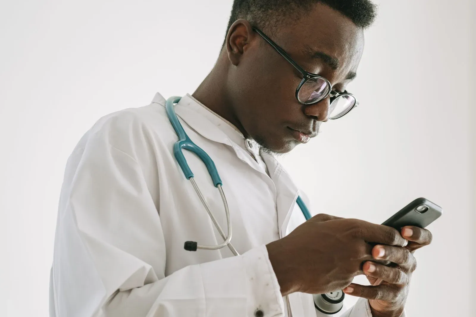 A health worker in a white coat with a stethoscope reads a message on his phone