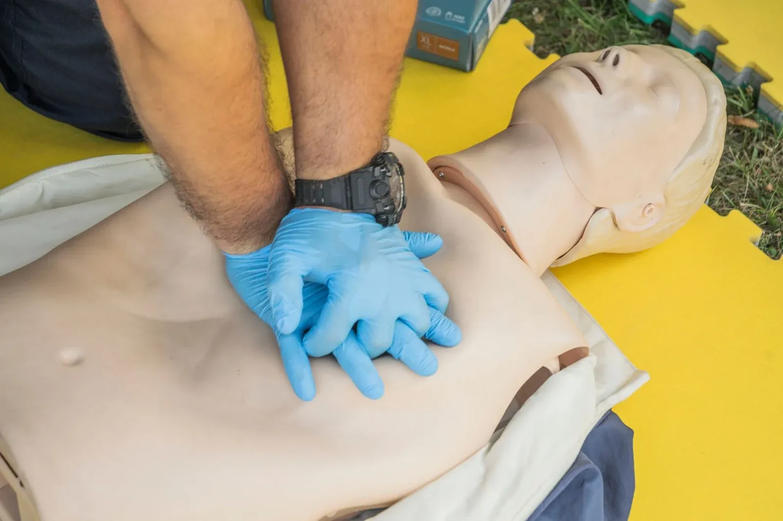 Gloved hands do chest compressions on an adult training manikin lying on a yellow mat